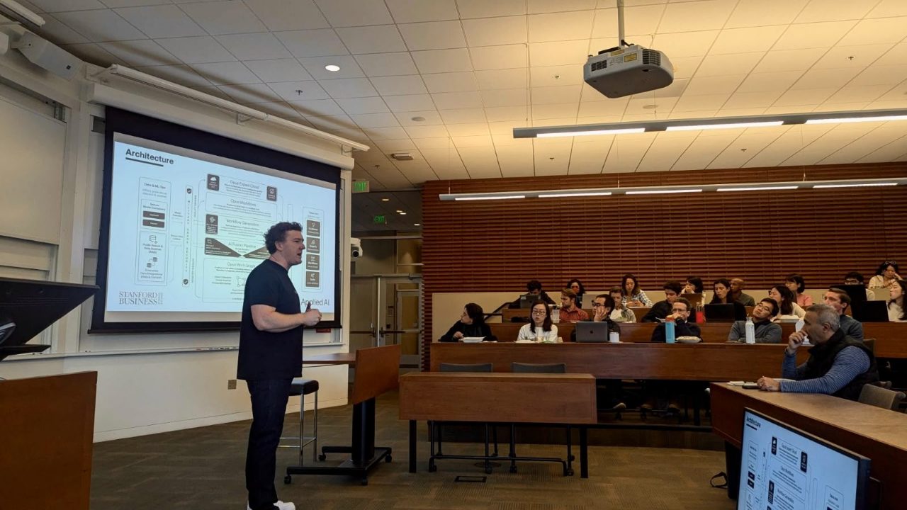 Phillip Kingston presenting the Opus architecture diagram to a tiered classroom at Stanford Graduate School of Business, the Stanford Business and AppliedAI marks at the foot of the slide.
